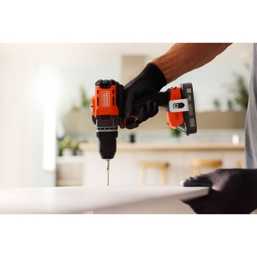 BLACK+DECKER cordless drill in use, drilling into a white surface indoors with a gloved hand.