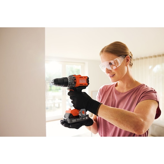 Person using a BLACK+DECKER cordless drill with safety glasses indoors, drilling into a wall.