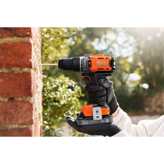 BLACK+DECKER cordless drill in use, drilling into a brick wall outdoors, held by a gloved hand.