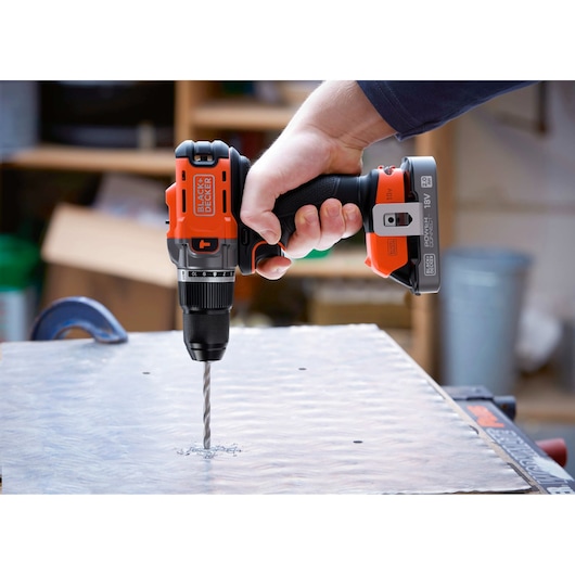 A BLACK+DECKER cordless drill is used to drill into a metal sheet on a workbench in a workshop.