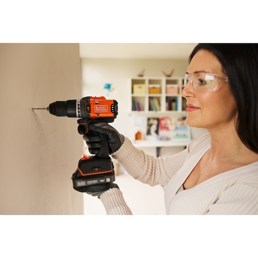 A woman drills into an interior wall using the BLACK+DECKER® 18V POWERCONNECT™ Brushless Hammer Drill.