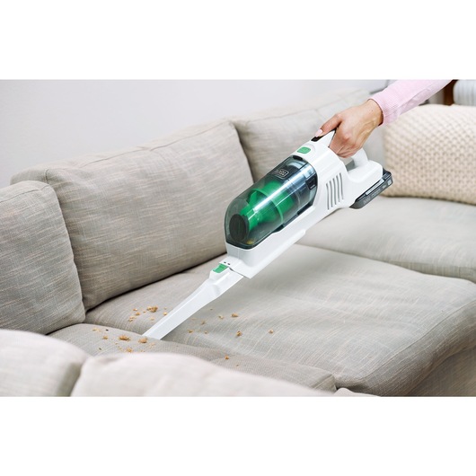 Crumbs on a sofa being vacuumed by the BLACK+DECKER® reviva™ 18V POWERCONNECT™ Cordless Stick Vacuum in handheld mode with crevice tool.